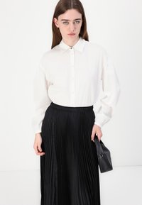 Baltā pogātā krekla ar garām piedurknēm kombinācija ar melnu plisētu svārku. Svārkiem ir elastīga josta un gluda tekstūra.