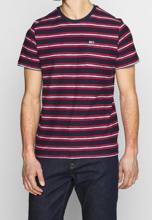 Mężczyzna ubrany w granatowy t-shirt z czerwono-białymi poziomymi paskami i ciemnoniebieskie dżinsy, stojący na jednolitym tle.
