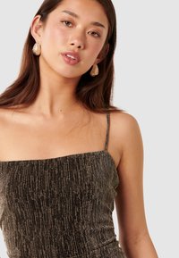 Robe noire avec des fils métalliques, présentant un décolleté carré et de fines bretelles. Le mannequin porte des boucles d'oreilles en or texturé.