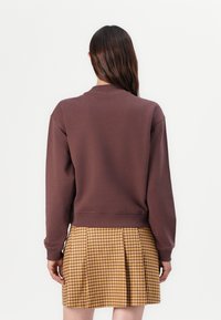 Brun genser med rund hals og lange ermer, brukt over et gult og brunt rutete miniskjørt med plissee. Jevn tekstur.