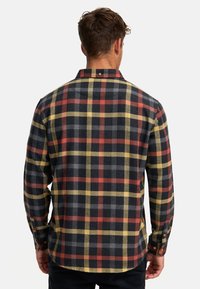 Homme portant une chemise à carreaux noire, rouge, jaune et grise à manches longues, vu de dos sur un fond blanc uni.