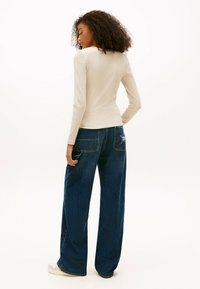 Mujer con cabello rizado que lleva un top ajustado de manga larga beige y unos jeans oscuros holgados con un parche de estrella en el bolsillo trasero.