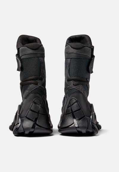 Svarta ankelstövlar med ett nätövre, texturerade stödpaneler, justerbara detaljer och en gummiaktig sula med distinkta mönster.