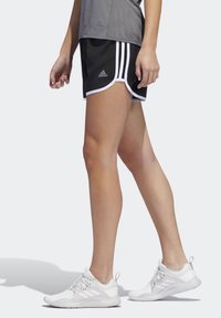 Shorts deportivos negros con ribetes blancos, que presentan tres rayas blancas a los lados. Combinados con zapatillas blancas y una camiseta gris.