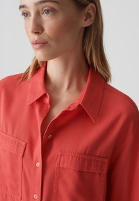 Camicia button-up color corallo con colletto, dotata di due tasche sul petto e una texture liscia, indossata leggermente aperta sul collo.