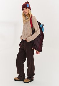 Béžový svetr s copánkovým vzorem, hnědé kalhoty s širokými nohavicemi, bordó čepice s modrým štítkem, batoh s bordó a modrými detaily, hnědé šněrovací boty.