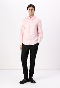 Camisa de botones rosa claro, pantalones slim fit negros y botas de cuero negras. El modelo lleva gafas rectangulares. Tejido suave, ajuste a medida.
