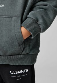 Felpa grigia con polsini a coste e una tasca frontale, abbinata a pantaloni neri con scritte del marchio bianche. Tessuto morbido e strutturato.