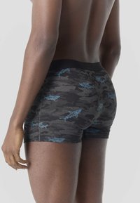 Primer plano de una persona vistiendo pantalones cortos de camuflaje negro y gris con patrones de tiburones azules, mostrada de espaldas desde la cintura hacia abajo.