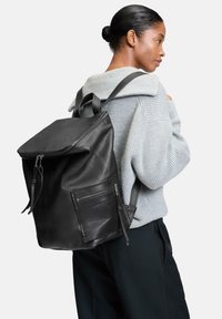 Sac à dos en cuir noir avec un dessus zipp é, finition texturée et une poche latérale zippée. Porté sur un pull gris.