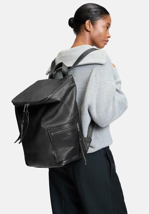 Schwarzer Leder-Rucksack mit einem Reißverschluss oben, strukturiertem Finish und einer seitlichen Tasche mit Reißverschluss. Über einem grauen Pullover getragen.