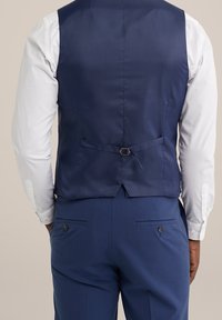 Námořnický modrý vest z hladké látky, s detailem s přezkou na zadní straně, kombinovaný se světlým šedým košilem a tmavě modrými kalhotami.