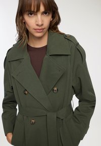Oliwkowy, dwurzędowy płaszcz trench z szerokimi klapami, ozdobnymi guzikami i paskiem do wiązania. Wykonany z gładkiego materiału, z wyraźnymi szwami.