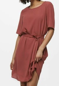 Mujer con un vestido rosa empolvado a la altura de la rodilla, con mangas cortas y un cinturón atado en la cintura, sosteniendo parte de la falda con una mano.