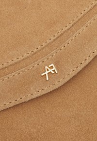 Tan mocka material med en mjuk textur. Har en guldfärgad metall-logotyp som accent och stickade kanter, vilket ger en sofistikerad och ren design.