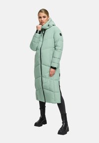 Woman wearing a long, light green padded coat with hood, black leggings, and black chunky boots, standing against a white background.
