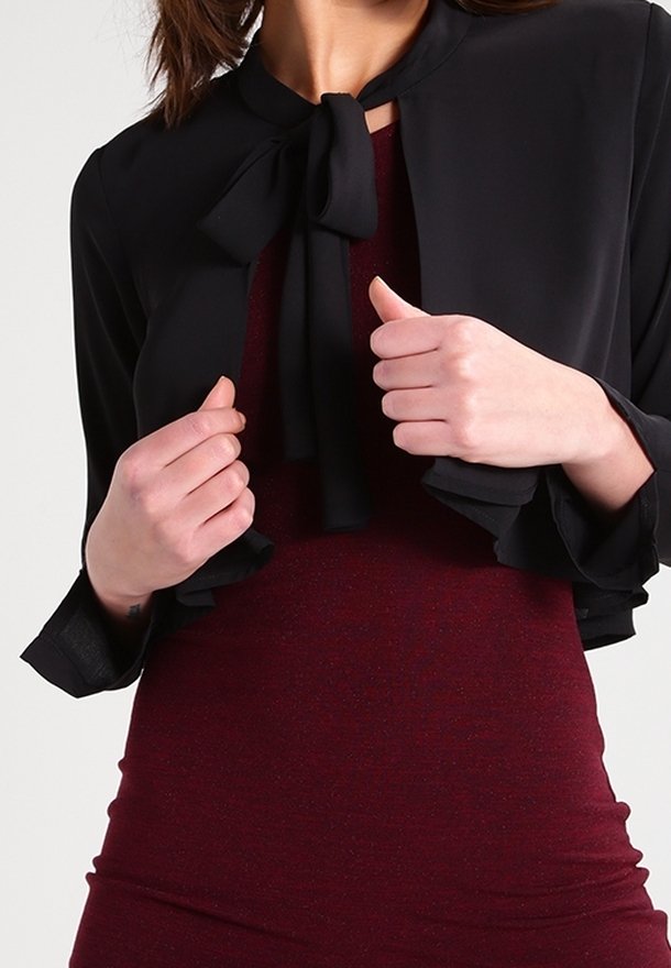 Femme portant une veste courte noire avec un nœud papillon sur une robe bordeaux ajustée, tenant le devant de la veste avec les deux mains.