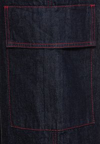 Mörkblå denimficka med lock, detaljerad med kontrasterande röda stygn vid kanterna och sömmarna.