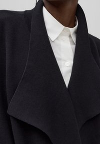 Musta villasekoitetakki, jossa leveä kaulus, kerroksellinen muotoilu ja teksturoitu kangas, joka laskeutuu valkoisen kauluspaidan päälle, jossa on nappidetaljit.