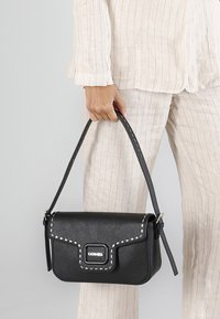 Svart handväska gjord av texturerat material, med silverfärgade nitar längs fliken och en axelrem. Rektangulär design med varumärkeslogotyp.