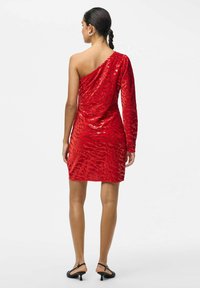 Raudona blizgučių suknelė su viena ilga rankove, prigludęs dizainas ir tekstūrinis raštas. Lygi medžiaga atspindi šviesą, suknelės ilgis iki kelių.
