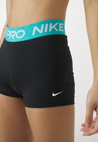 Gros plan d'une personne portant un short Nike Pro noir avec une ceinture turquoise affichant le logo "NIKE PRO" et un logo Nike Swoosh blanc.