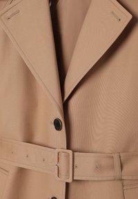 Gros plan sur un trench-coat beige avec de larges revers, des boutons noirs et une ceinture assortie avec une boucle rectangulaire.