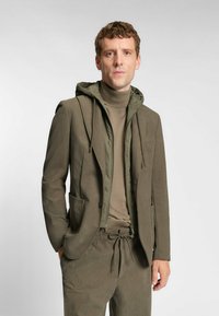 Olijfgroen pak met een met een capuchon afgewerkte ritsjack, in laag over een beige coltrui. Bevat een zak, een aantrekkoord in de taille en een gestructureerde stof.