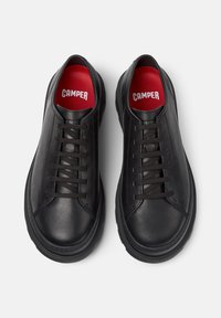 Un paio di sneaker nere in pelle con lacci e suole spesse, con soletti rossi del marchio "Camper", visti dall'alto su uno sfondo bianco.