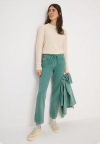 Jeans de algodón verde con un corte relajado, combinados con un suéter texturizado en beige claro y zapatillas beige. Se sostiene una chaqueta a juego en una mano.
