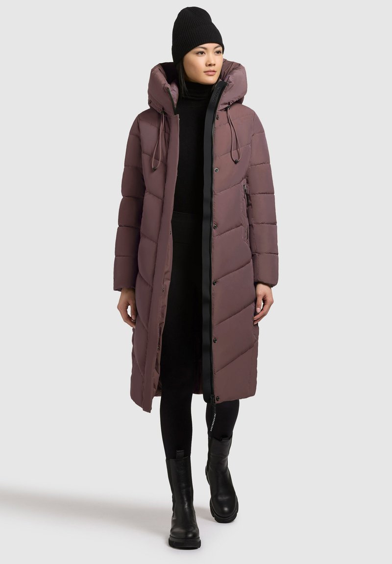 Lang pufferjakke i dæmpet mauve med lodret quiltet mønster, hætte med snøre og sort kant. Bæres sammen med sort tætsiddende tøj og støvler.