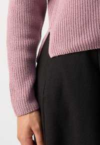 Różowy sweter z dzianiny o prążkowanej fakturze i bocznych rozporkach, noszony z czarnymi spodniami o fakturze, podkreślając kontrast między materiałami.