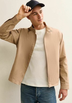 Joven ajustándose la gorra negra, vistiendo una chaqueta beige con cremallera, una camiseta blanca y jeans azules, de pie contra un fondo claro y liso.