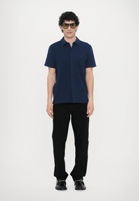 Navy short-sleeve button-up shirt with a subtle logo, paired with black trousers and black shoes featuring decorative hardware accents.
