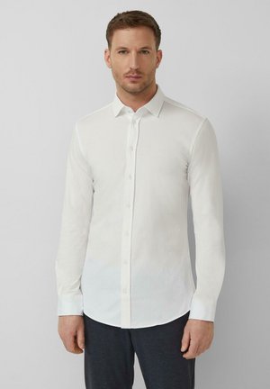 Mężczyzna ubrany w dopasowaną białą koszulę na guziki z długimi rękawami i kołnierzykiem, stojący na jednolitym szarym tle.