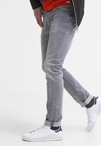 Šedé slim-fit džíny s jemnou texturou, s ohrnutými nohavicemi, spárované s bílými teniskami s námořnickými akcenty. Oblečený v červené košili a černé bundě.