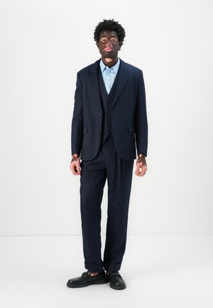Man with vitiligo wearing a navy suit, light blue shirt, and black loafers standing against a plain white background.