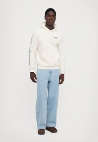 Cream hoodie with drawstring, front pocket, and EA7 logo. Paired with light blue wide-leg jeans and brown footwear. Plain background.