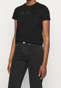 Černé bavlněné tričko s krátkým rukávem, které je zdobeno texturovaným designem z kamínků na hrudi. V kombinaci s černými džíny.