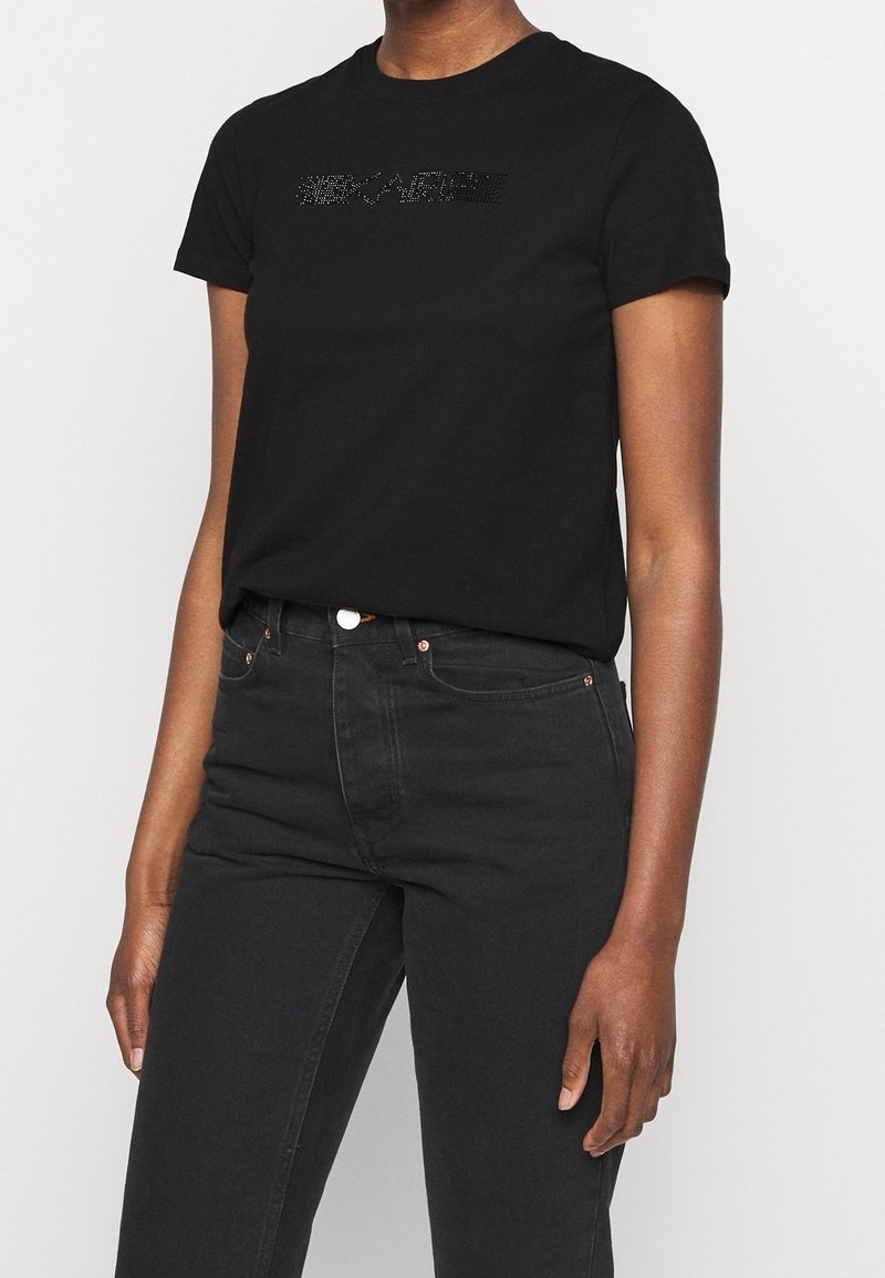 Černé bavlněné tričko s krátkým rukávem, které je zdobeno texturovaným designem z kamínků na hrudi. V kombinaci s černými džíny.