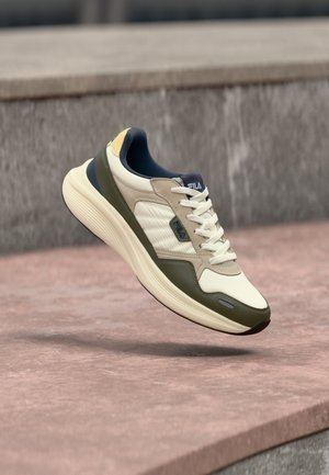 Fehér és olívazöld FILA sportcipő vastag talppal, bézs és tengerészkék díszítéssel, kövekkel borított járda felett lebegve, beton párkány közelében.