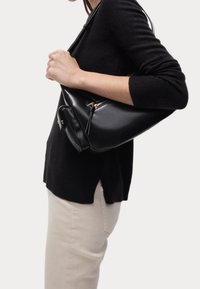 Donna che indossa un maglione nero e pantaloni beige, mentre porta una borsa a spalla in pelle nera con cerniera e una piccola tasca attaccata.
