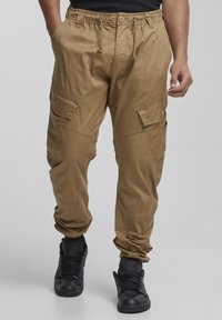 Bež cargo hlače z elastičnim pasom, več žepov in nastavljivimi manšetami. Nošene s črnimi supergami, ki imajo gladko teksturo.