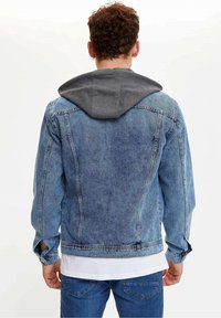 Giubbotto in denim di colore azzurro chiaro con cappuccio grigio. Le caratteristiche includono una vestibilità comoda, tasche laterali e cuciture visibili. Vista posteriore mostrata.