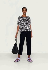 Top preto com padrão floral de mangas curtas, calças largas pretas, sandálias lavanda e uma bolsa tote preta texturizada, tudo em contraste com um fundo neutro.
