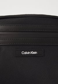 Zaino nero con superficie in tessuto testurizzato, dettagli lisci, una tasca con zip e un'etichetta con il logo "Calvin Klein" bianco.