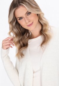 Cardigan lavorato a maglia color crema con polsini a coste e apertura frontale, indossato sopra una maglia beige chiaro. Texture morbida con una vestibilità rilassata.
