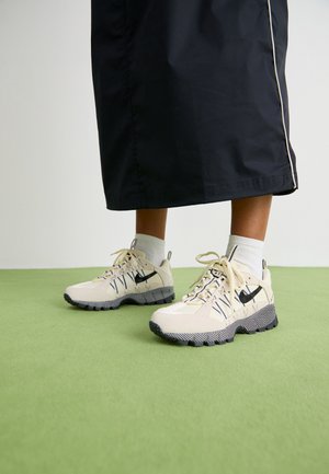 Person iført beige Nike sneakers med sorte såler og hvide sokker, stående på grøn gulv, iført en lang sort nederdel.