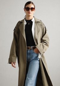 Trench coat verde oliva con solapas anchas, usado sobre una camiseta negra y una camisa blanca. Jeans de mezclilla y cinturón marrón completan el conjunto.