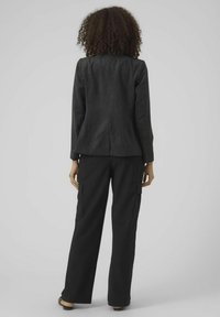 Schwarzer Cordblazer mit taillierter Passform, kombiniert mit lockeren schwarzen Cargo-Hosen mit seitlichen Taschen. Der Hintergrund ist schlicht grau.